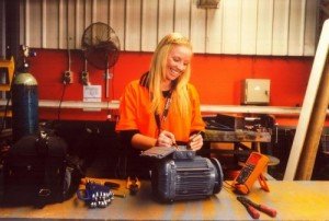 A young woman in a bright orange smock is at a workbench, smiling as she uses equipment to test an electrical system.
